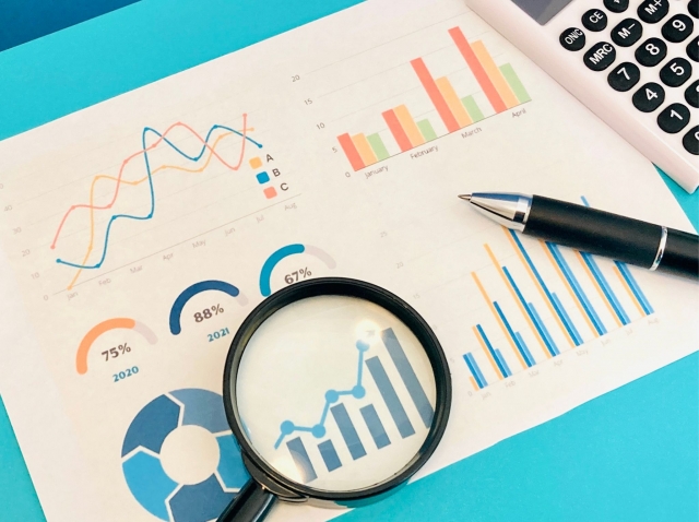 様々なグラフが印刷された紙の上に、虫眼鏡とボールペンが置かれている画像